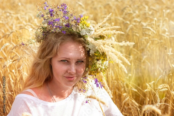 Fototapeta Beautiful blonde with long hair and a wreath of flowers on her head sits in a field of ripe wheat.