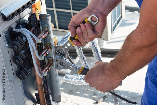 Fototapeta HVAC technician is working on air conditioner units on a roof of new industrial building.Technician hand using fix wrench to tighten outdoor unit of air condition. Man holds a wrench in his hand.
