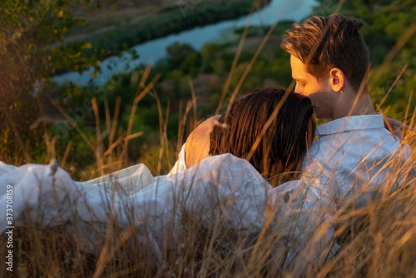 Fototapeta half portrait of a beautiful couple of guy and girl lying in the grass. Against the backdrop of the setting sun on a summer evening. White dress of the bride.