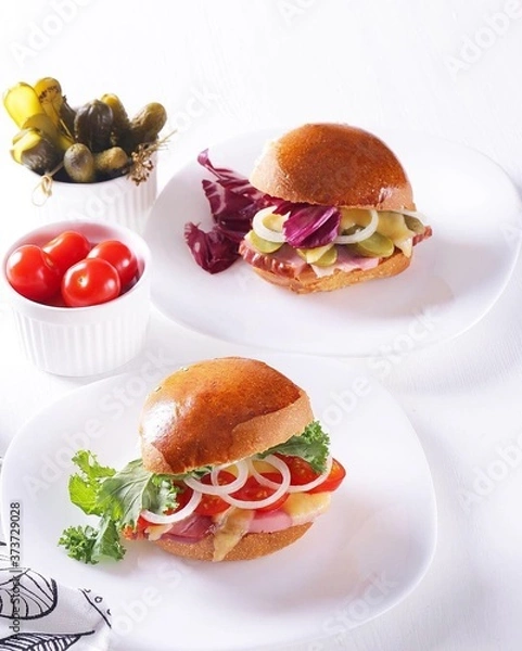 Fototapeta on a white table on white plates are two hamburgers with different fillings, next to cucumbers and tomatoes