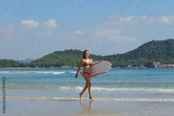 Fototapeta young slender and tanned girl in a swimsuit with a surfboard in her hands on the beach near the water, sea, summer, heat, sunny day, clear sea water, wave, lifestyle, sports, leisure, vacation, weeken