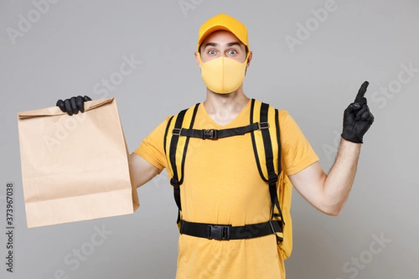 Fototapeta Delivery employee man in yellow cap face mask gloves tshirt thermal backpack hold craft paper takeaway food bag work courier service during quarantine covid-19 virus isolated on grey background studio