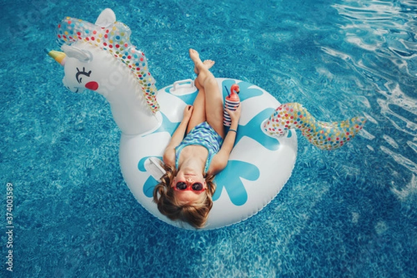 Fototapeta Cute adorable girl in sunglasses with drink lying on inflatable ring unicorn. Kid child enjoying having fun in swimming pool. Summer outdoors water activity for kids. View from above overhead.