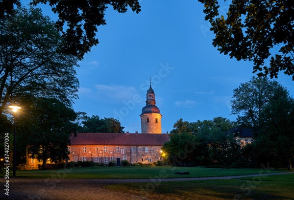 Obraz illuminated medieval former monastery and tower of the church St. Viti in Zeven (Germany) seen through large trees in the city park during scenic blue hour at dawn