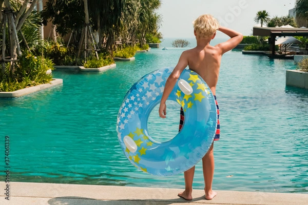 Fototapeta Happy little boy standing near the swimming pool with a blue and yellow pool float in his hands. Copy space banner.