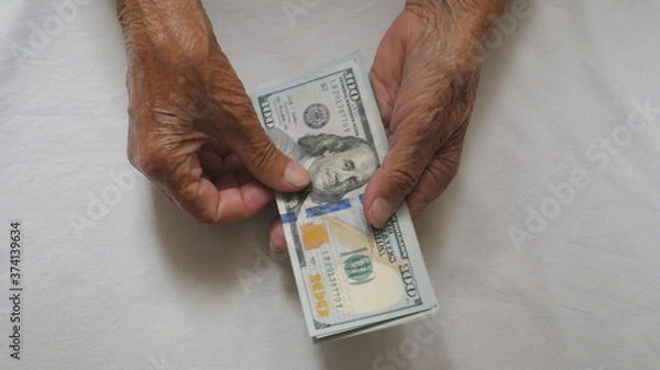 Fototapeta Wrinkled female hands holds cash and counts foreign currency over the table. Arms of elderly grandmother puts one hundred dollar banknotes on the desk. Money concept. Top view Slow motion
