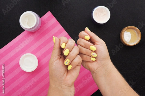 Obraz female hands with bright yellow manicure. on the black table there are jars of cream, varnish and a pink napkin..