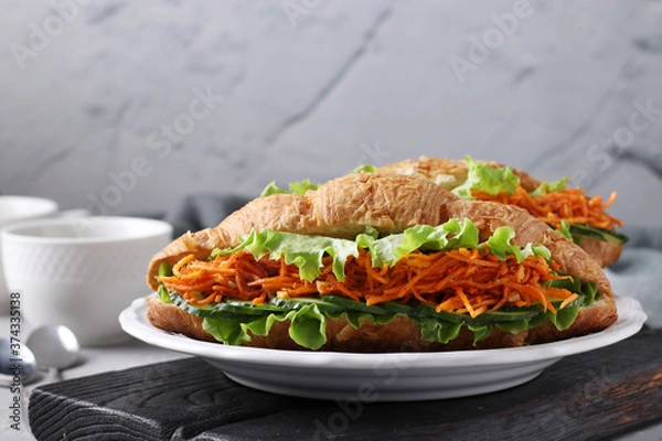 Fototapeta Two vegetarian croissants-sandwiches with lettuce, carrots, cucumbers and two cups of coffee on a light grey concrete background. Breakfast or lunch concept. Closeup