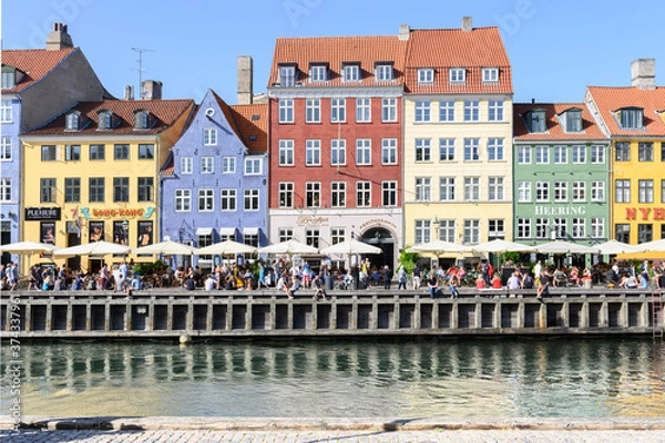 Fototapeta COPENHAGEN, DENMARK 29. June 2019: People relax on a promenade along Nyhavn in front of a row of colorful houses on a warm summer's day.