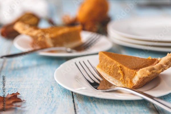 Fototapeta Zbliżenie kawałek ciasta dyniowego na talerzu widelcem z talerzami i innymi przedmiotami rozmazanymi w tle na niebiesko malowanej rustykalnej drewnianej powierzchni stołu