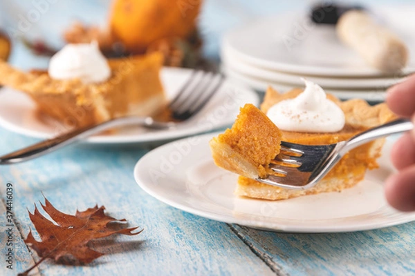 Fototapeta Closeup perspective shot of a bite of pumpkin pie on a fork with plates and slices of pumpkin pie blurred in the background on a rustic blue painted wood table surface.