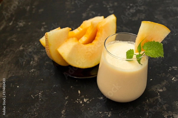 Fototapeta Melon milk shake on a black background. Ripe sliced melon is lying nearby. The smoothie glass is decorated with a sprig of mint and melon crumbs. Summer healthy food concept