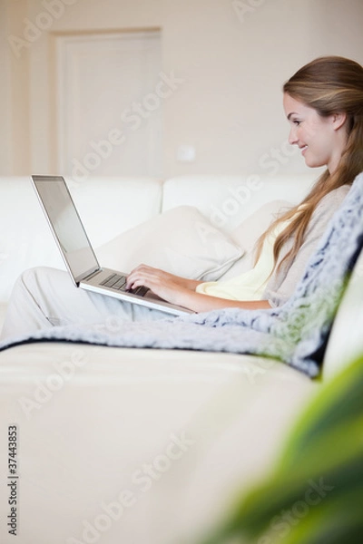 Fototapeta Side view of woman with her laptop sitting on the sofa