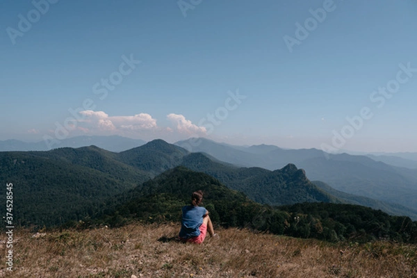 Fototapeta Beautiful mountain and hilly landscape in warm, Sunny weather and the young tourist. A female traveler sits on top of a hill and looks at the surrounding mountains.