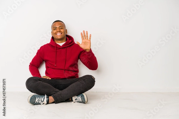 Fototapeta Young latin man sitting on the floor isolated smiling cheerful showing number five with fingers.