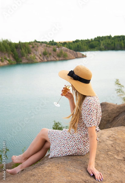 Fototapeta A girl with blond hair sits on a mountain above the lake and looks down.