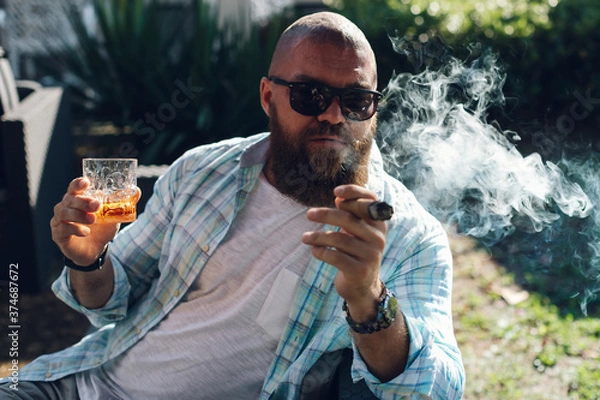Obraz Portrait of an attractive bearded business man smoking a cigar with glass of whiskey in a summer cafe.