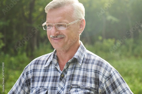 Fototapeta Close-up portrait of handsome gray-haired adult man with mustache and glasses smiling on the background of a summer forest park