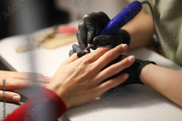 Fototapeta Professional in black gloves make mechanized manicure to visitor in studio. Woman processes her nails before applying varnish in workplace.