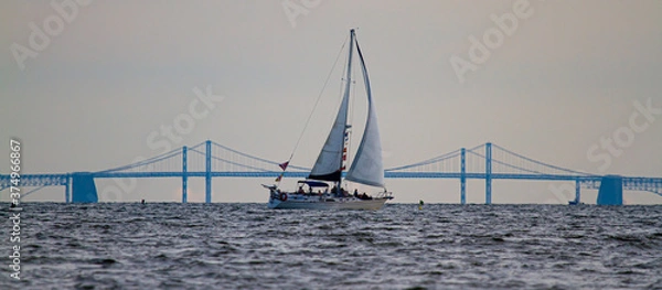 Fototapeta Panoramiczny obraz żaglówki poruszającej się po zatoce Chesapeake z sylwetką słynnego mostu Bay Bridge w tle. Na łodzi są ludzie, którzy podziwiają zachód słońca nad zatoką.