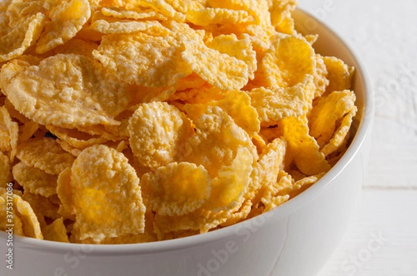 Fototapeta White bowl of cornflakes on a white wooden table on the left side of the frame. .Close-up.