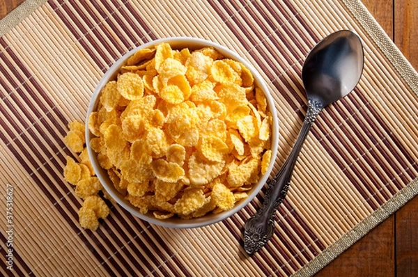 Fototapeta A white bowl of cornflakes, next to a metal spoon, all on a napkin that is on a brown wooden table. Composition in the center of the frame. Horizontal frame.