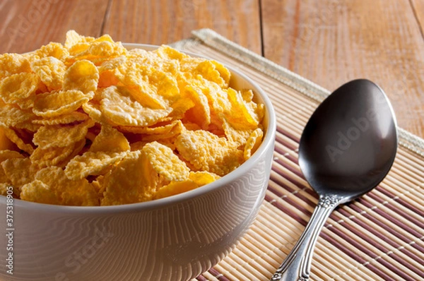 Fototapeta A white bowl of cornflakes, next to a metal spoon, all on a napkin that is on a brown wooden table. Horizontal frame.