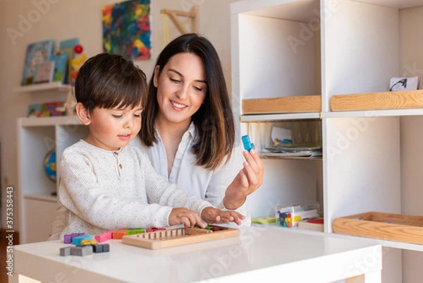 Obraz Kid playing with a tetris wood puzzle and mother or teacher help. Homeshooling. Learning Community. Montessori School
