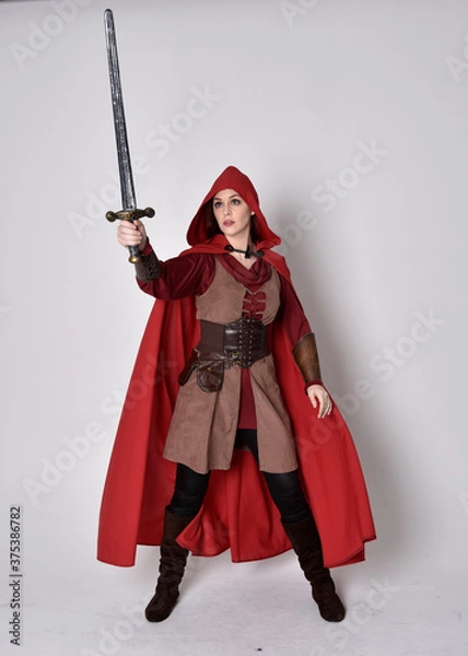Fototapeta Full length portrait of girl wearing medieval costume and red cloak. Standing pose holding a sword,  isolated against a grey studio background.