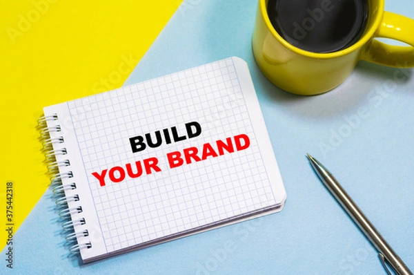 Obraz BUILD YOUR BRAND on a notepad on a blue-yellow background, a cup of coffee and a pen. Business concept