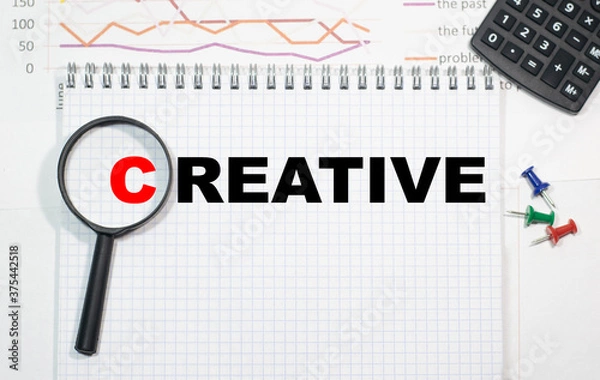 Obraz CREATIVE in a magnifying glass on a notepad against the background of graphs, calculator and paper clips.
