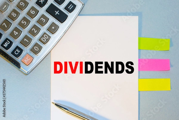 Obraz DIVIDENDS is written on a notepad next to multicolored stickers, a calculator and a pen on a blue background. Business concept