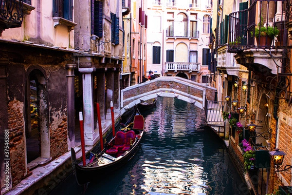 Obraz Beautiful Venice canal with gondola and red brick bridge. The luxurious gondola has red velvet-lined seats. On one wall there are lit lights and purple flowers in the balconies.