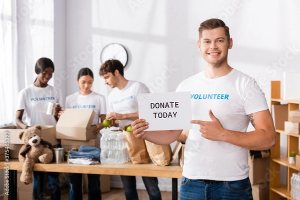 Fototapeta Selective focus of volunteer pointing at card with donate today lettering in charity center