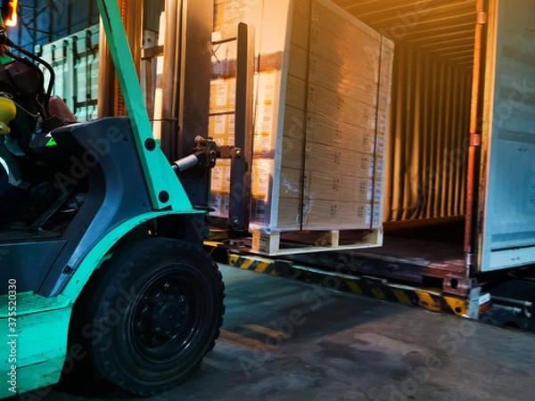 Fototapeta Worker driving forklift loading shipment carton boxes goods on wooden pallet at loading dock from container truck to warehouse cargo storage in freight logistics, transportation industrial, delivery