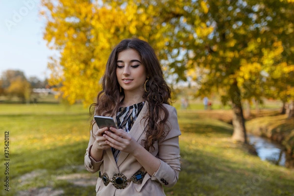 Fototapeta Beauty portrait of a cheerful girl with curly brunette hair in elegant suit reading on cell phone, in the park.
