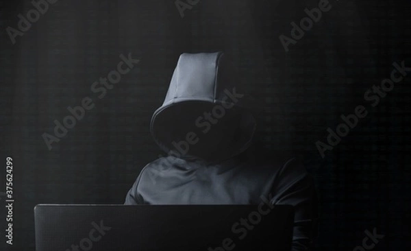 Fototapeta Hacker in a gray hood with a laptop on a dark background.