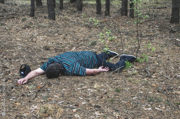 Obraz Murder in the woods. The body of a man in a blue t-shirt and trousers lies on the ground among the trees in the forest. Victim of an attack.