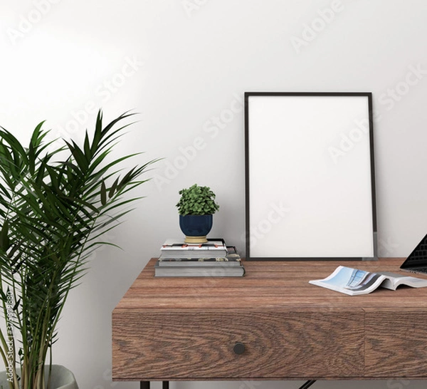 Fototapeta Vertical black frame above a walnut wood desk with books with a small blue vase with plants to one side, a magazine and a palm houseplant in a concrete vase to the side in front of a white wall