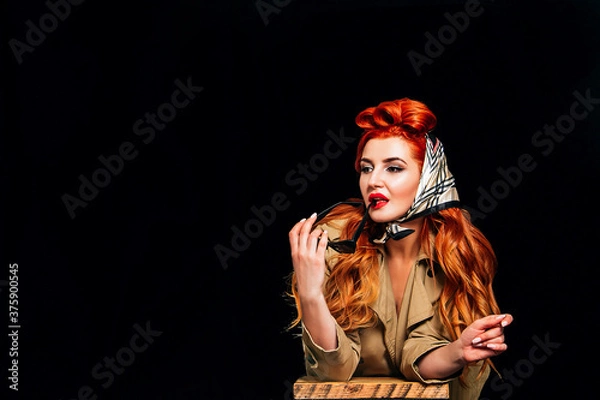 Fototapeta A beautiful red-haired girl on a black background dressed in the style of pin-up with red lipstick holds glasses and does not look at the camera. Valentine's Day, March 8, International Women's Day.
