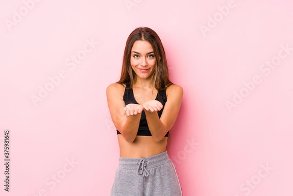 Fototapeta Young caucasian fitness woman posing in a pink background holding something with palms, offering to camera.