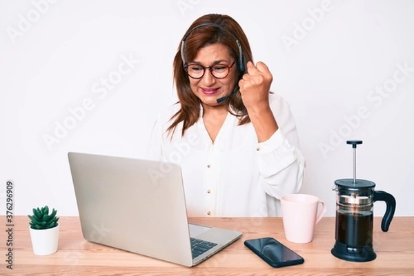 Fototapeta Middle age brunette hispanic woman working at the office wearing operator headset annoyed and frustrated shouting with anger, yelling crazy with anger and hand raised