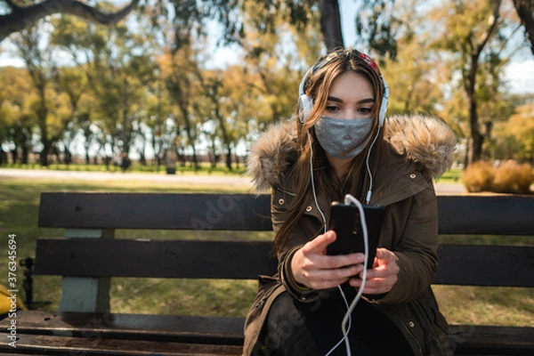 Fototapeta A caucasian woman wearing face mask sitting on a bench and using her smartphone. Copy space