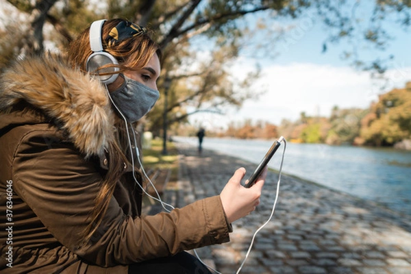 Fototapeta A caucasian woman wearing face mask and headphones relaxing and using her smartphone at park. Copy space