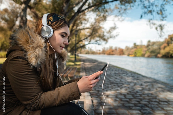 Fototapeta A beautiful woman wearing headphones and using her smartphone in front of a lake at park. Copy space