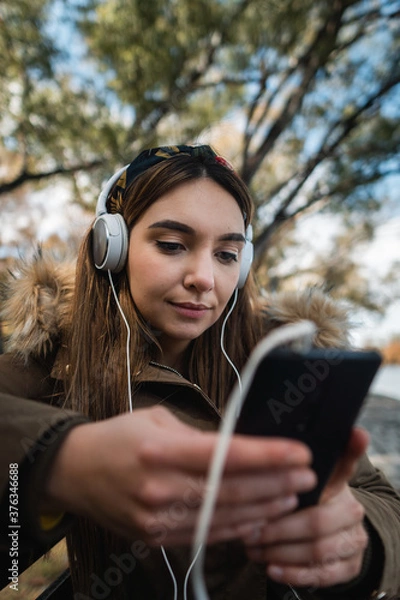Fototapeta Close up of a caucasian woman using headphones and texting with her smartphone at park