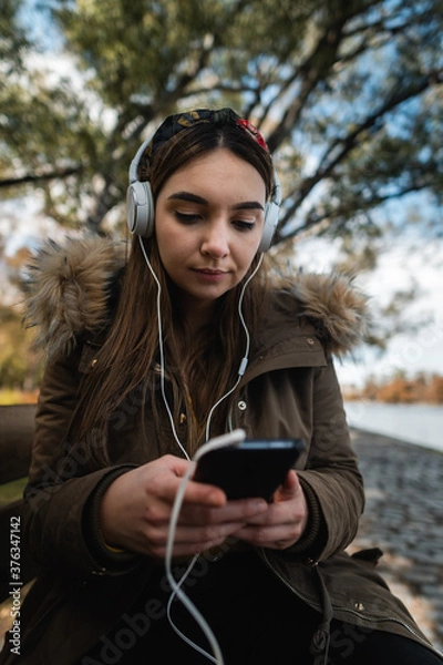 Fototapeta A caucasian woman using headphones and texting with her smartphone at park