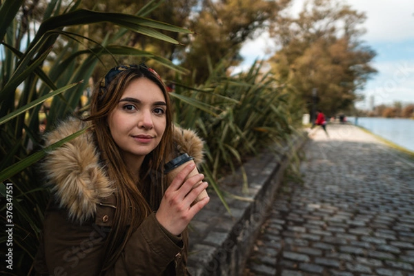 Fototapeta A pretty woman smiling at camera while relaxing at park and drinking a coffee. Vertical