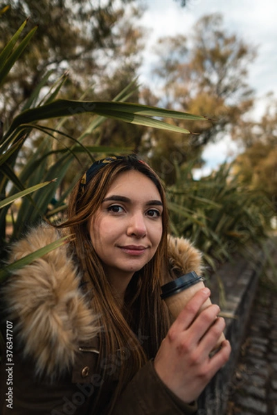 Fototapeta A pretty woman smiling at camera while relaxing at park and drinking a coffee. Vertical