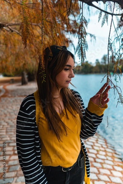 Fototapeta Vertical shot of a young woman touching a hanging leaf at autumnal park with a lake